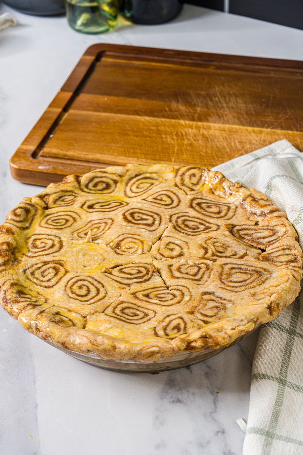 A pie dish with unbaked apple pie with cinnamon roll crust. The dish is on a marble counter with a wooden board.