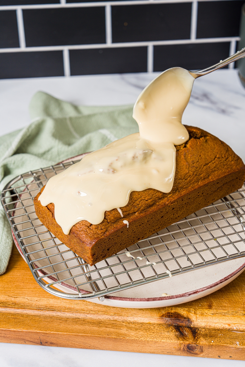 A wire rack over a white plate with a pumpkin loaf. A spoon is pouring maple glaze over the loaf. The plate is on a wooden board.