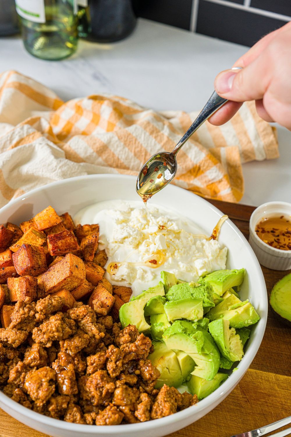 A white bowl with seasoned ground turkey, roasted sweet potatoes, avocado, and greek yogurt. A spoon is pouring hot honey over the yogurt.