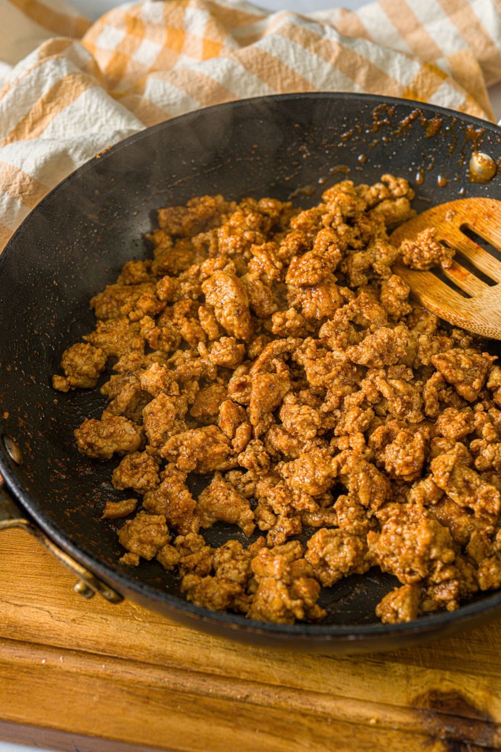 A skillet with a wooden spoon breaking up seasoned ground turkey. The skillet is on a wooden board with a yellow checkered napkin.