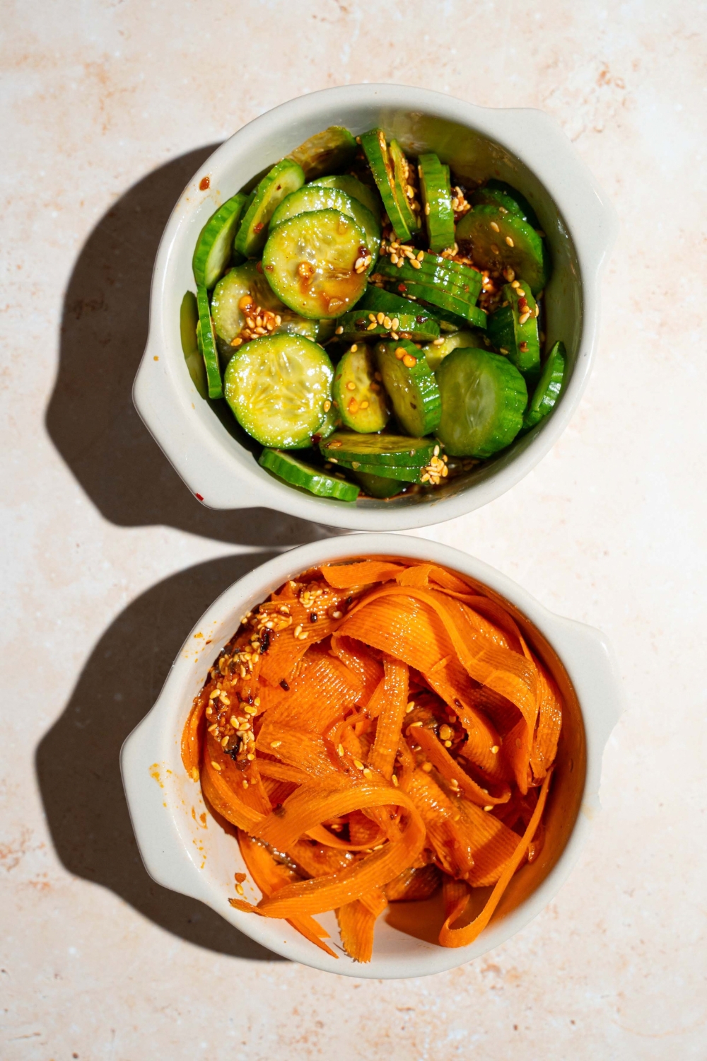 Two bowls of thinly sliced cucumbers and thinly sliced carrots tossed in a sesame soy sauce dressing. The bowls are on a tan counter.