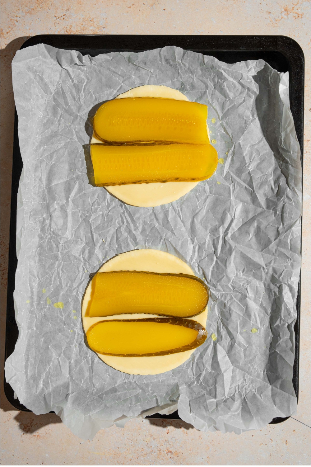 A baking sheet lined with parchment paper with two slices of cheese topped with sliced pickles.