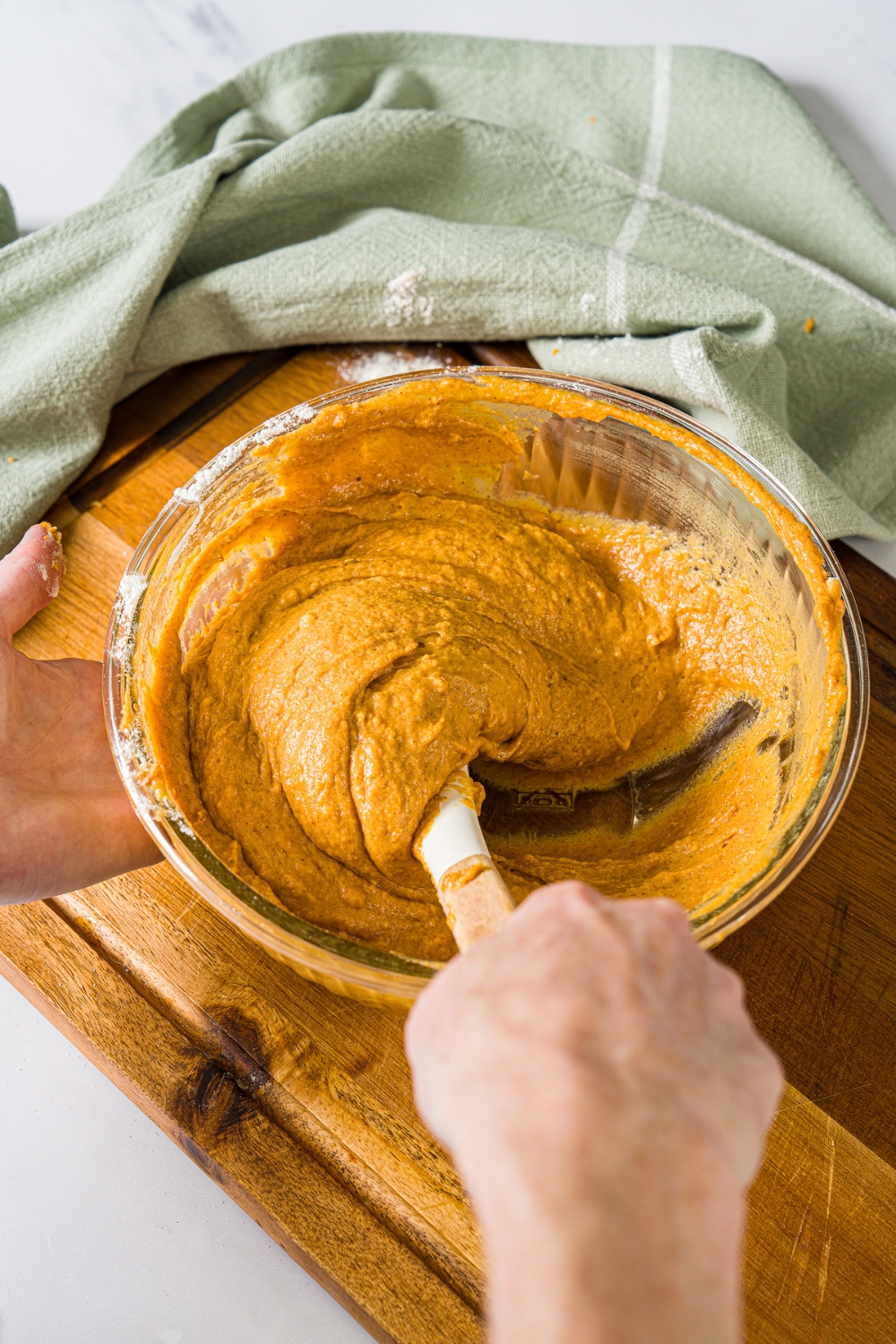 A large bowl with a spatula mixing pumpkin bread batter. The bowl is on a wooden board with a green cloth napkin.