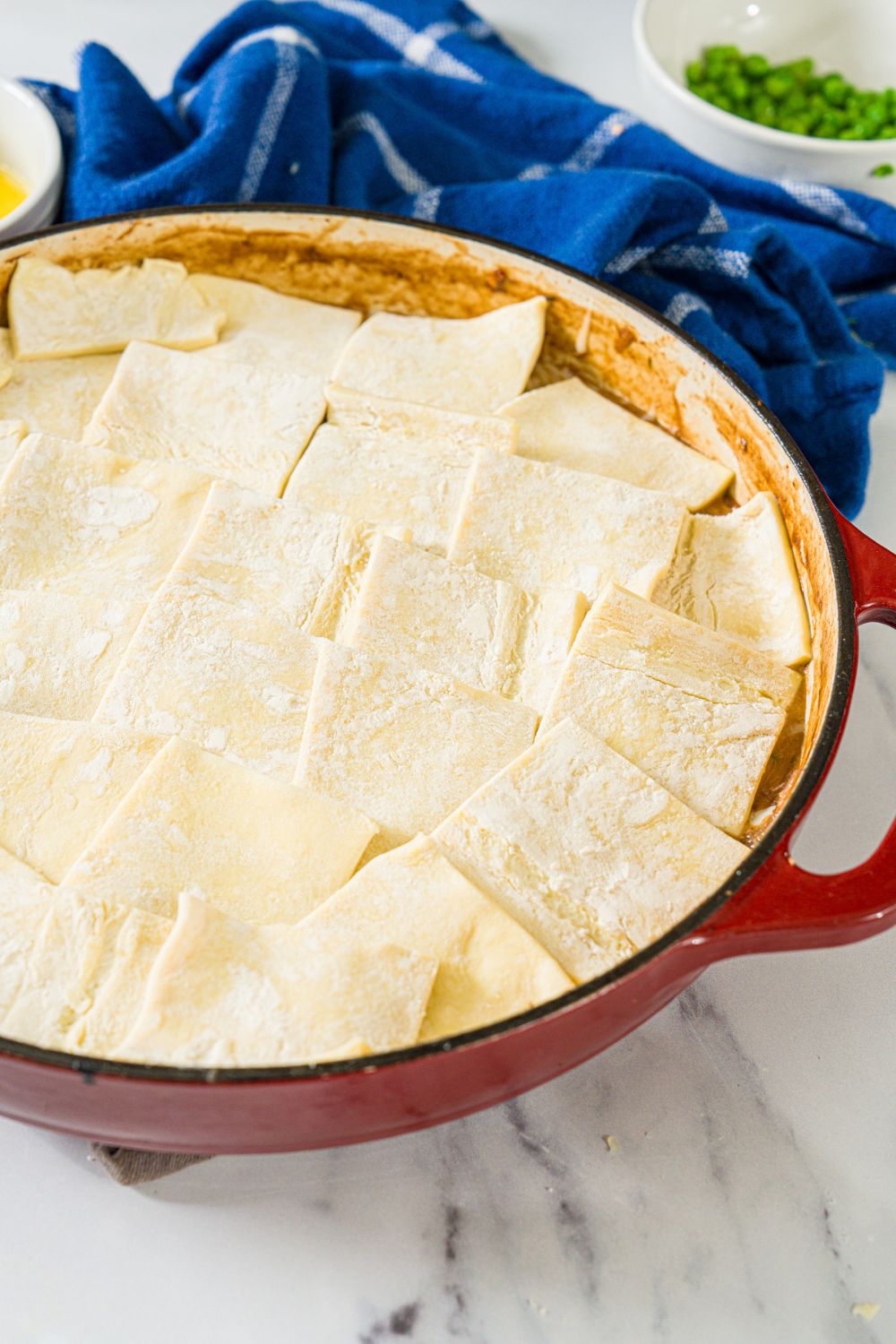 A dutch oven with a french onion chicken pot pie mixture topped with puff pastry squares. The dutch oven is on a marble counter with a blue cloth napkin.