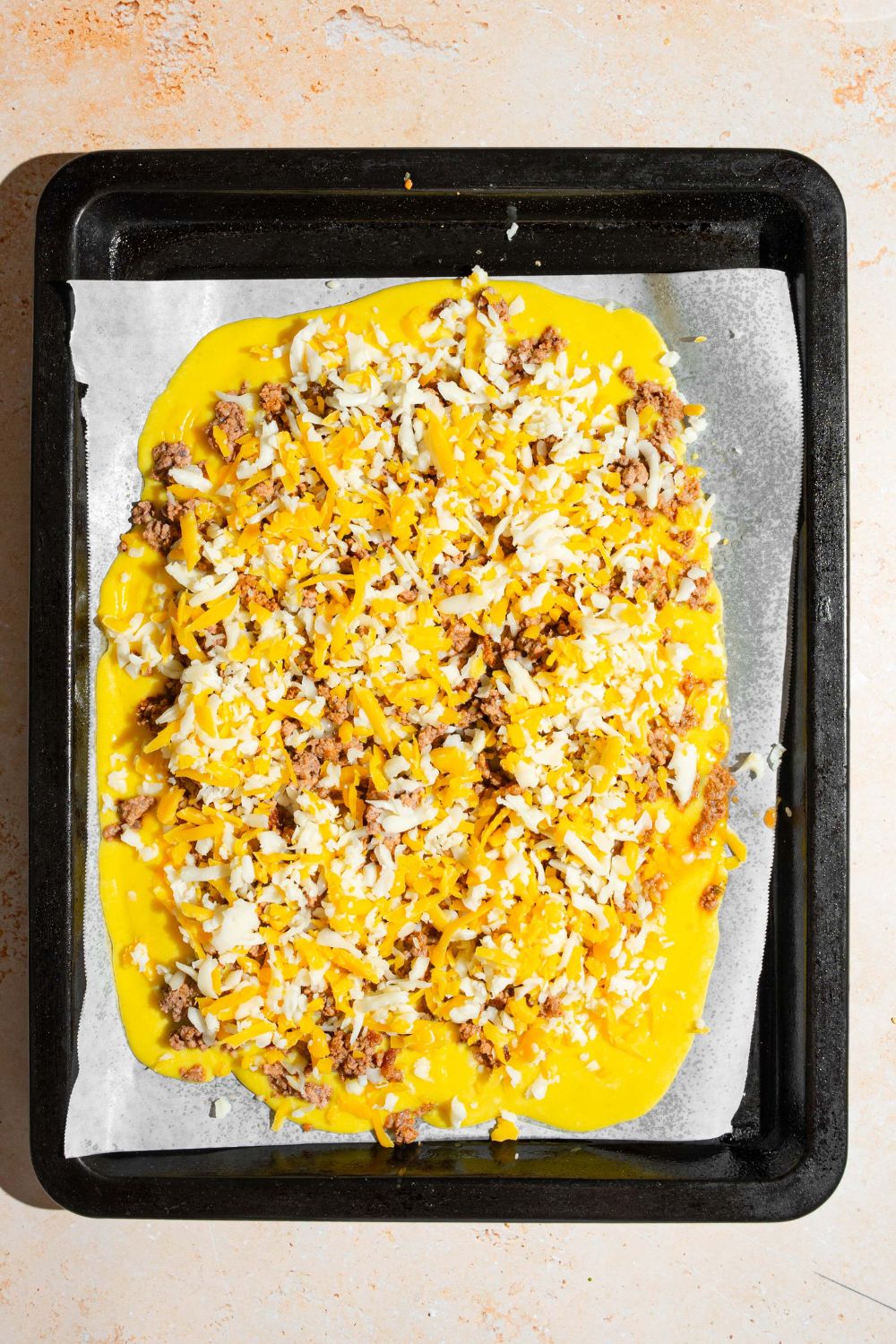 A baking tray lined with parchment paper with nacho cheese, seasoned ground beef, and shredded cheese on top. The tray is on a tan counter.