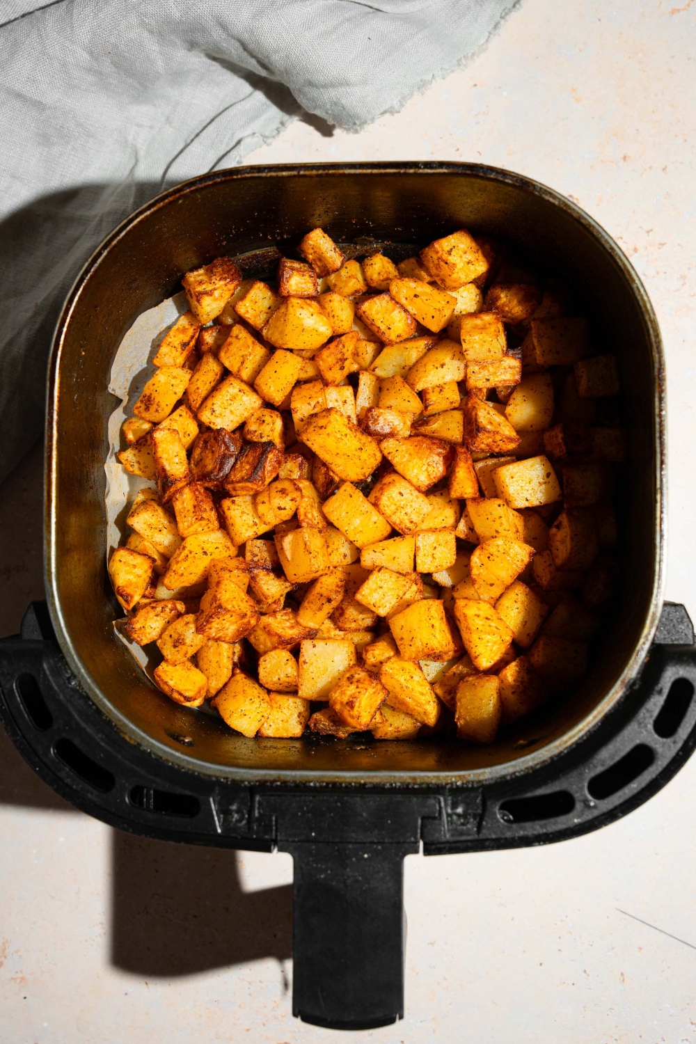 An air fryer basket with air fried seasoned potatoes. The basket is on a tan counter.
