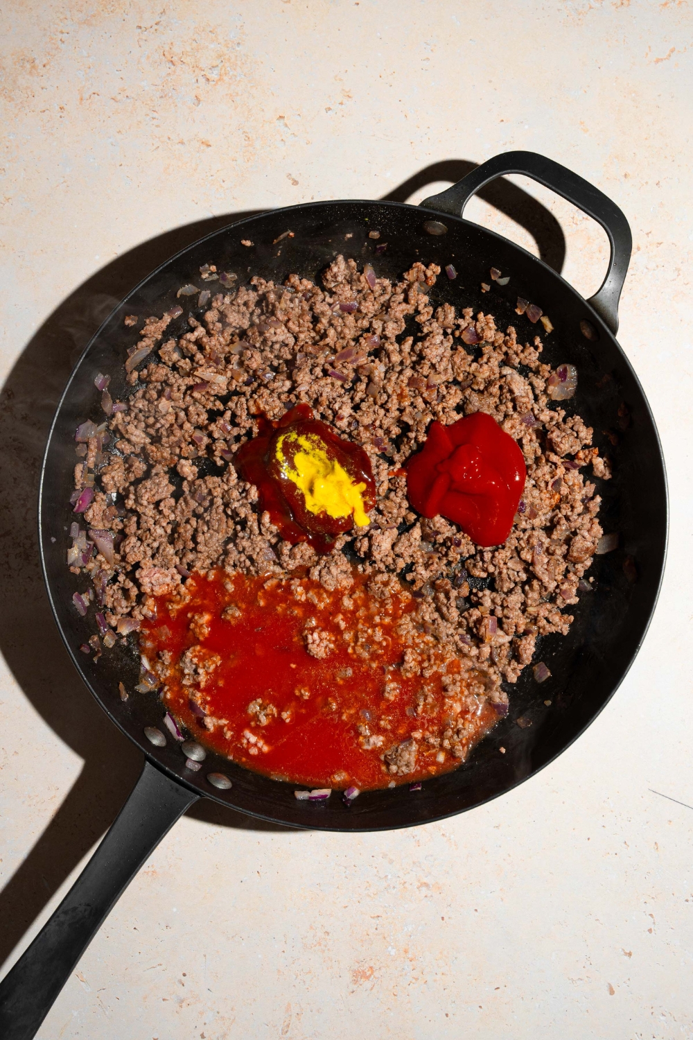 A skillet with ground beef mixture topped with ingredients to make sloppy joe sauce including ketchup, mustard, tomato sauce, and more. The skillet is on a tan counter.