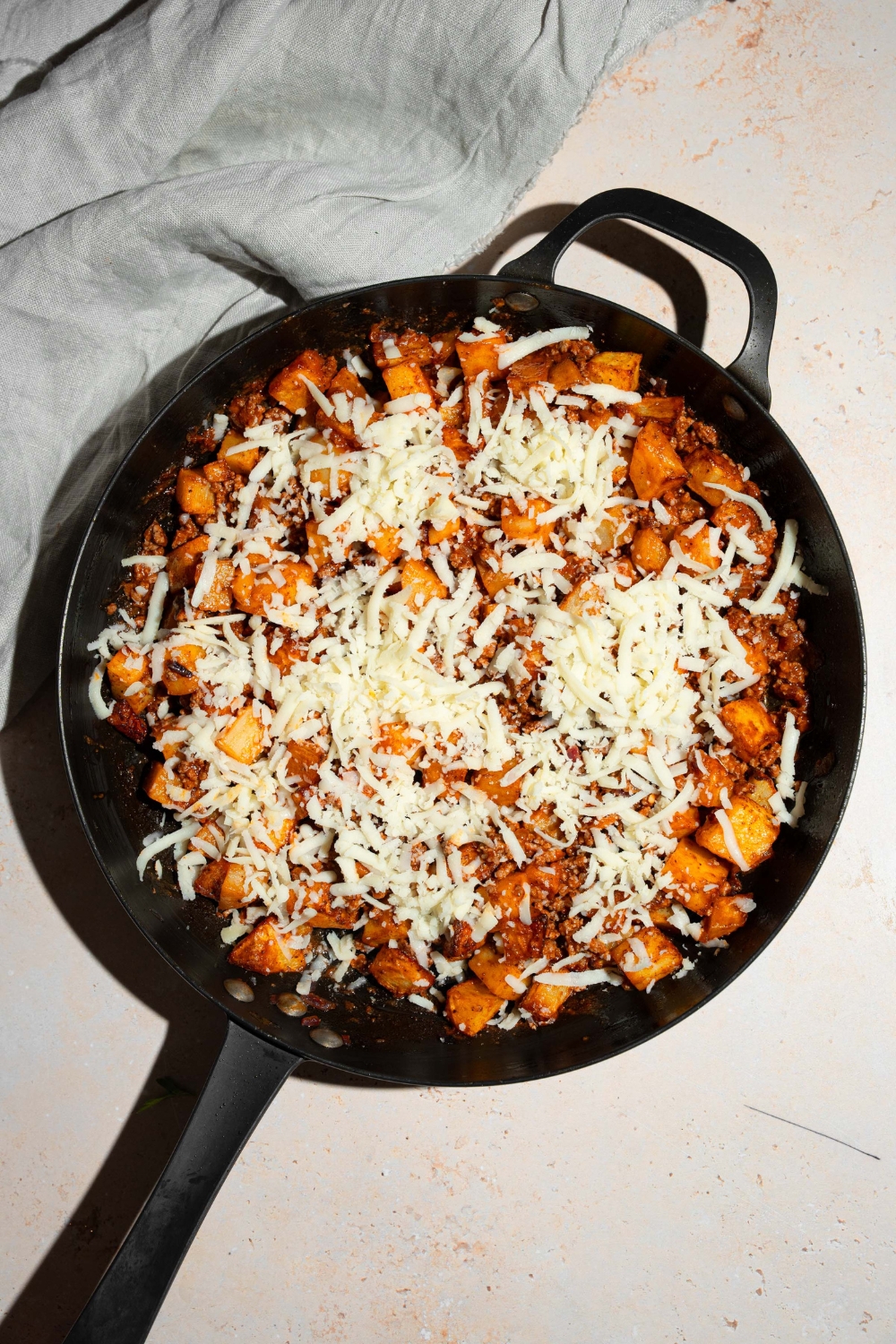 A skillet with a sloppy joe potato mixture topped with shredded cheese. The skillet is on a tan counter.