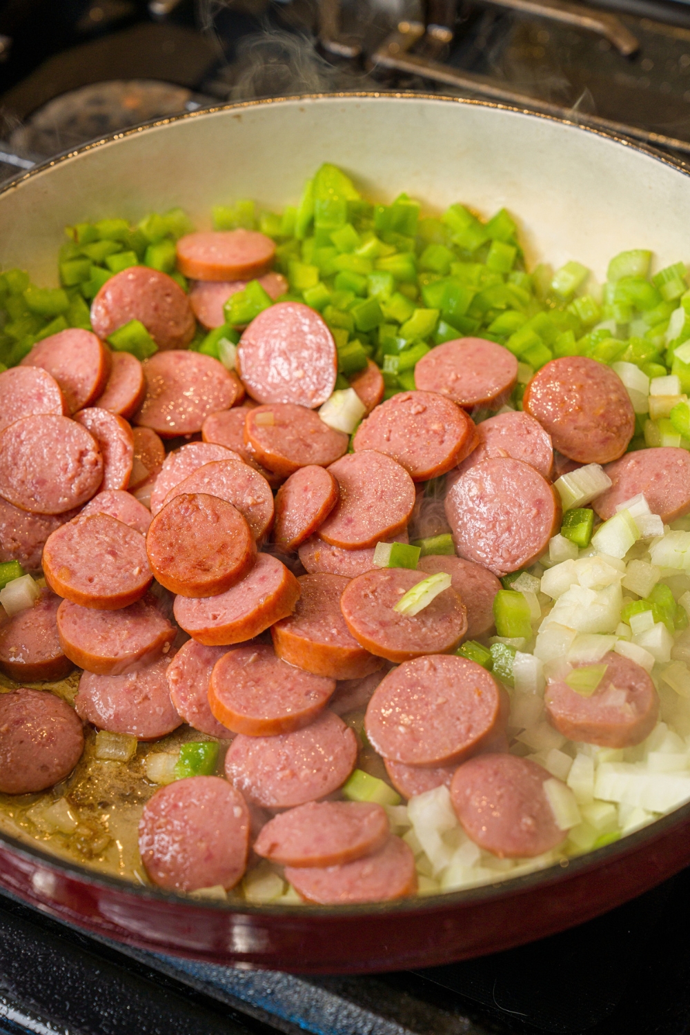 A skillet with sautéed celery and onion with sliced sausage. The skillet is cooking on a stove.