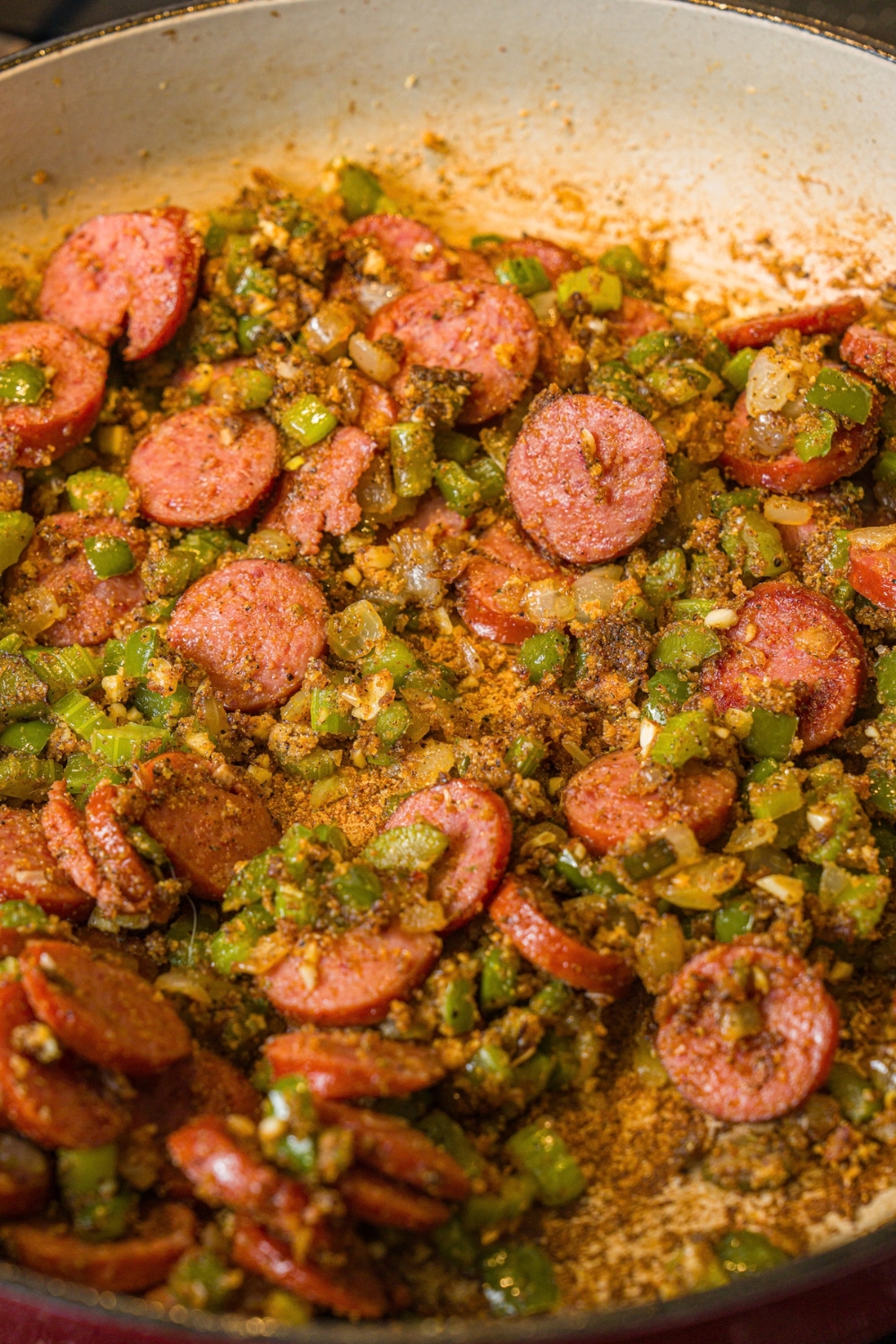 A skillet with sautéed sliced sausage and diced celery and onion. The skillet is cooking on a stove.