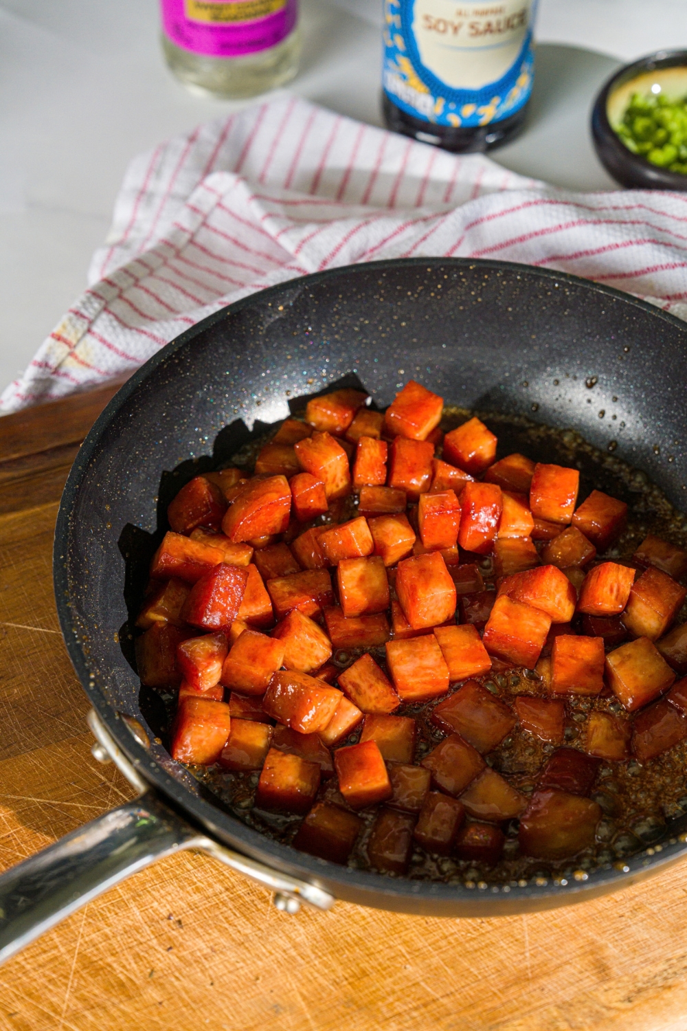 A skillet with spam sautéing in mirin soy sauce mixture. The skillet is on a wooden board.