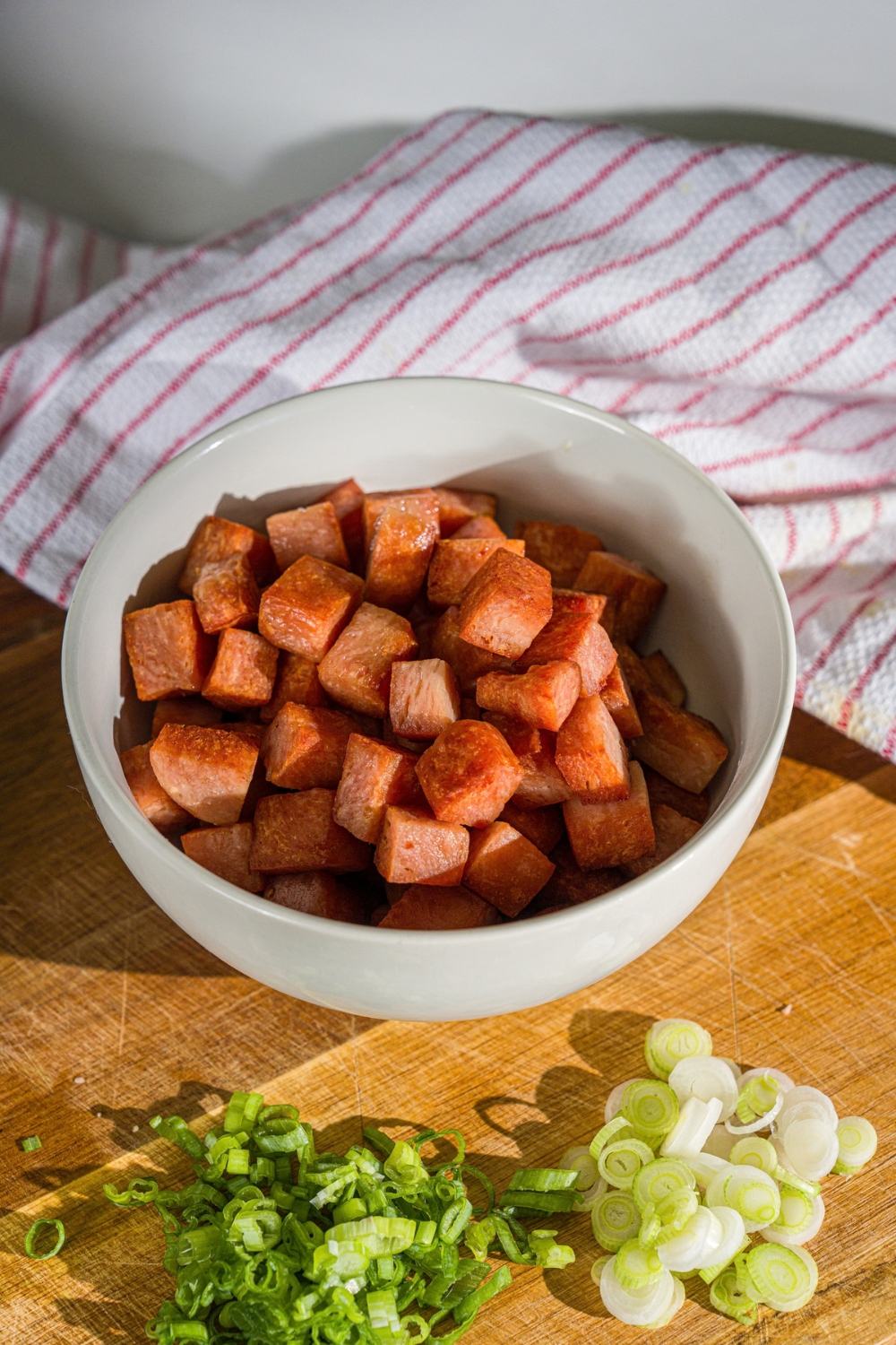 A small bowl with seared spam cubes. The bowl is on a wooden board with sliced green onion and a white cloth napkin.