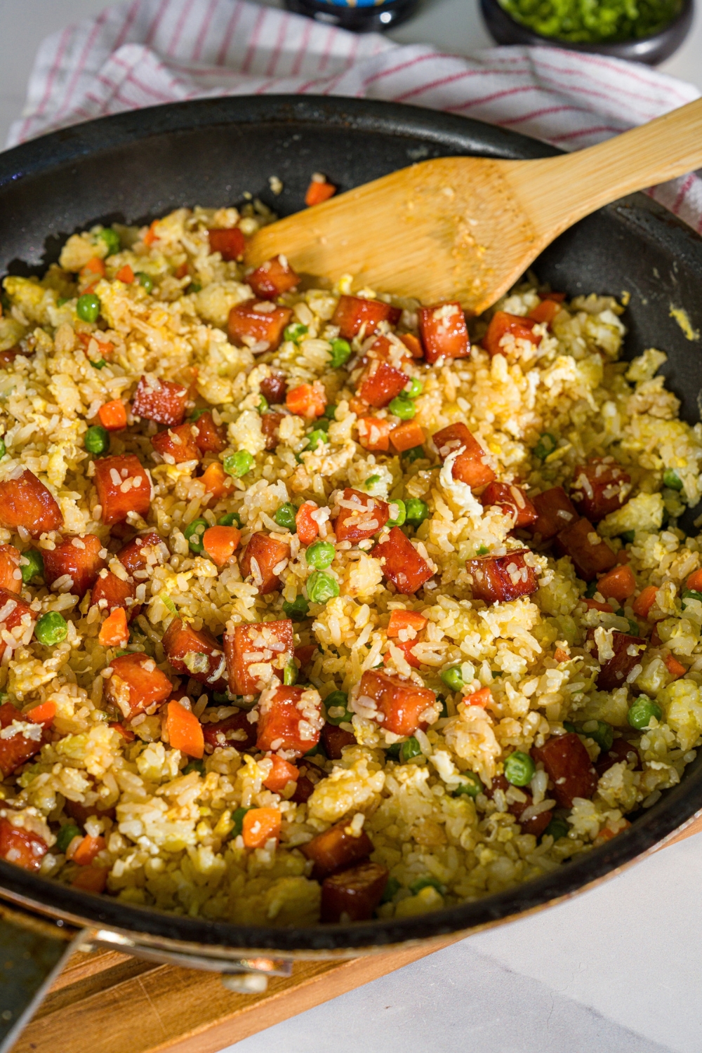 A skillet with a spam musubi rice mixture. There is a wooden spoon stirring the mixture. The skillet is on a wooden board.