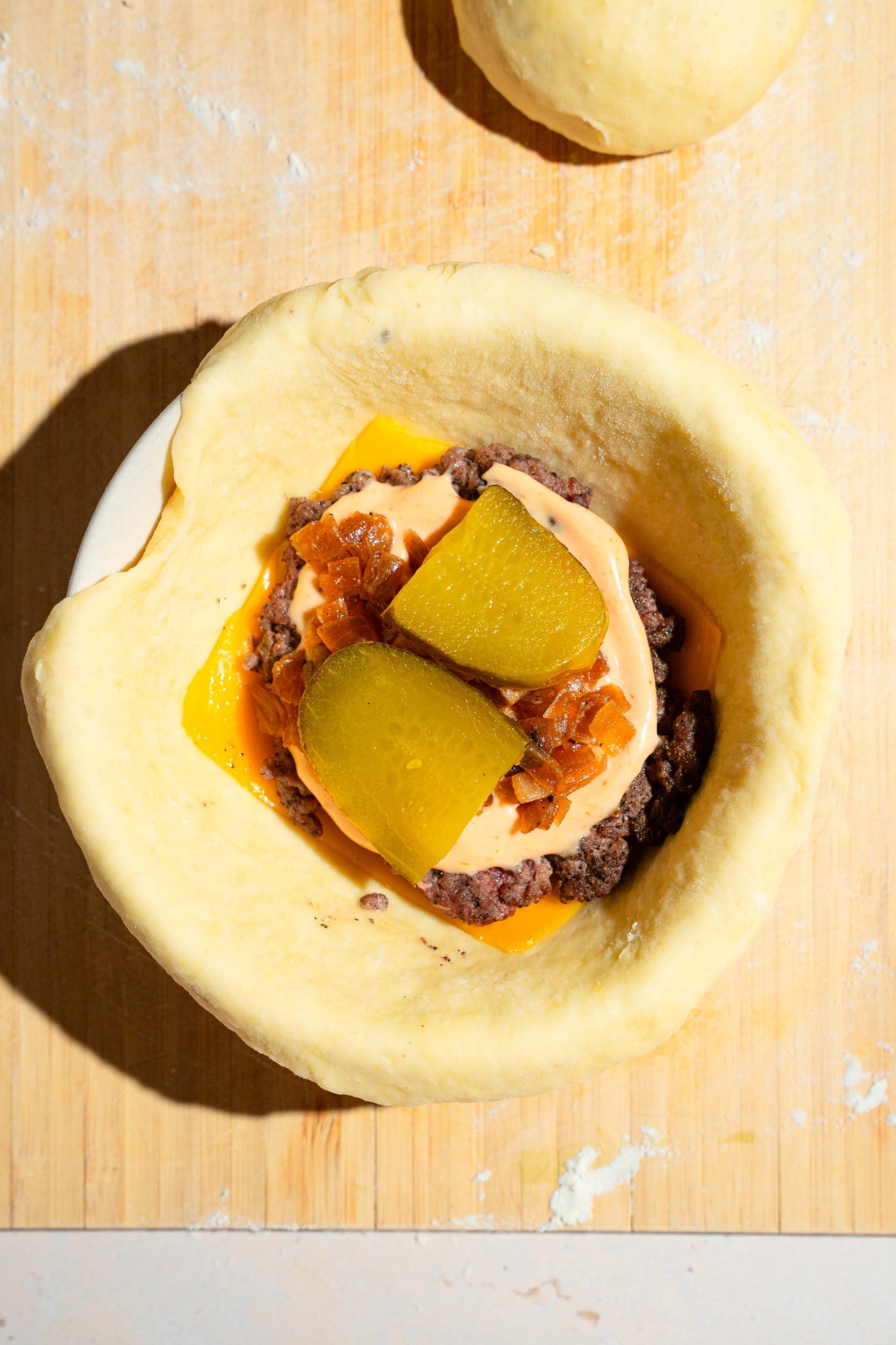 A small dough ball shaped in a ramekin with cheese a, smash burger patty, burger sauce, and pickles placed in the center.