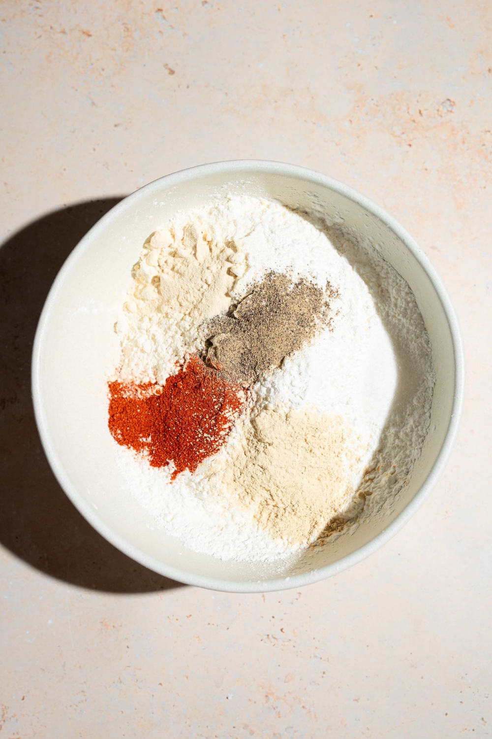 A large bowl with ingredients to make seasoned flour including flour, cornstarch, baking powder, garlic powder, smoked paprika, and more. The bowl is on a tan counter.