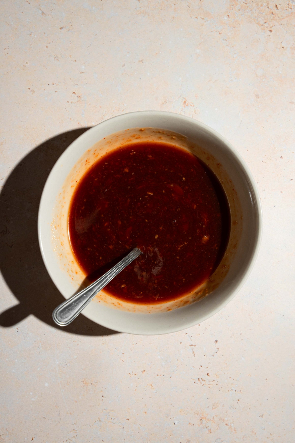 A small bowl with a sweet chili garlic sauce with a spoon in the bowl. The bowl is on a tan counter.
