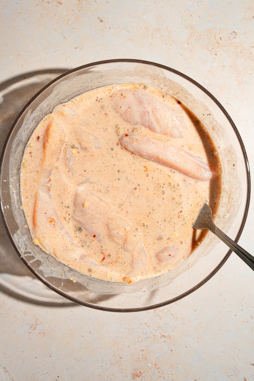 A large bowl with chicken tenders marinating in a wet batter mixture. The bowl is on a tan counter.