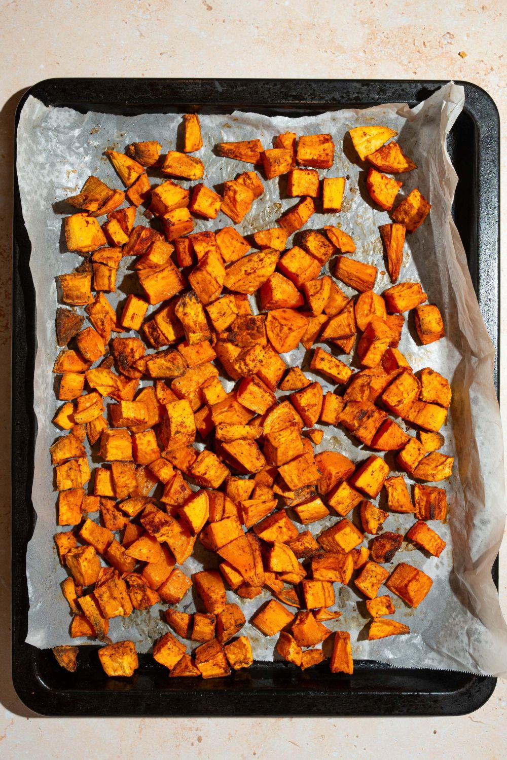 A baking sheet lined with parchment paper with roasted cubed seasoned sweet potatoes. The sheet is on a tan counter.