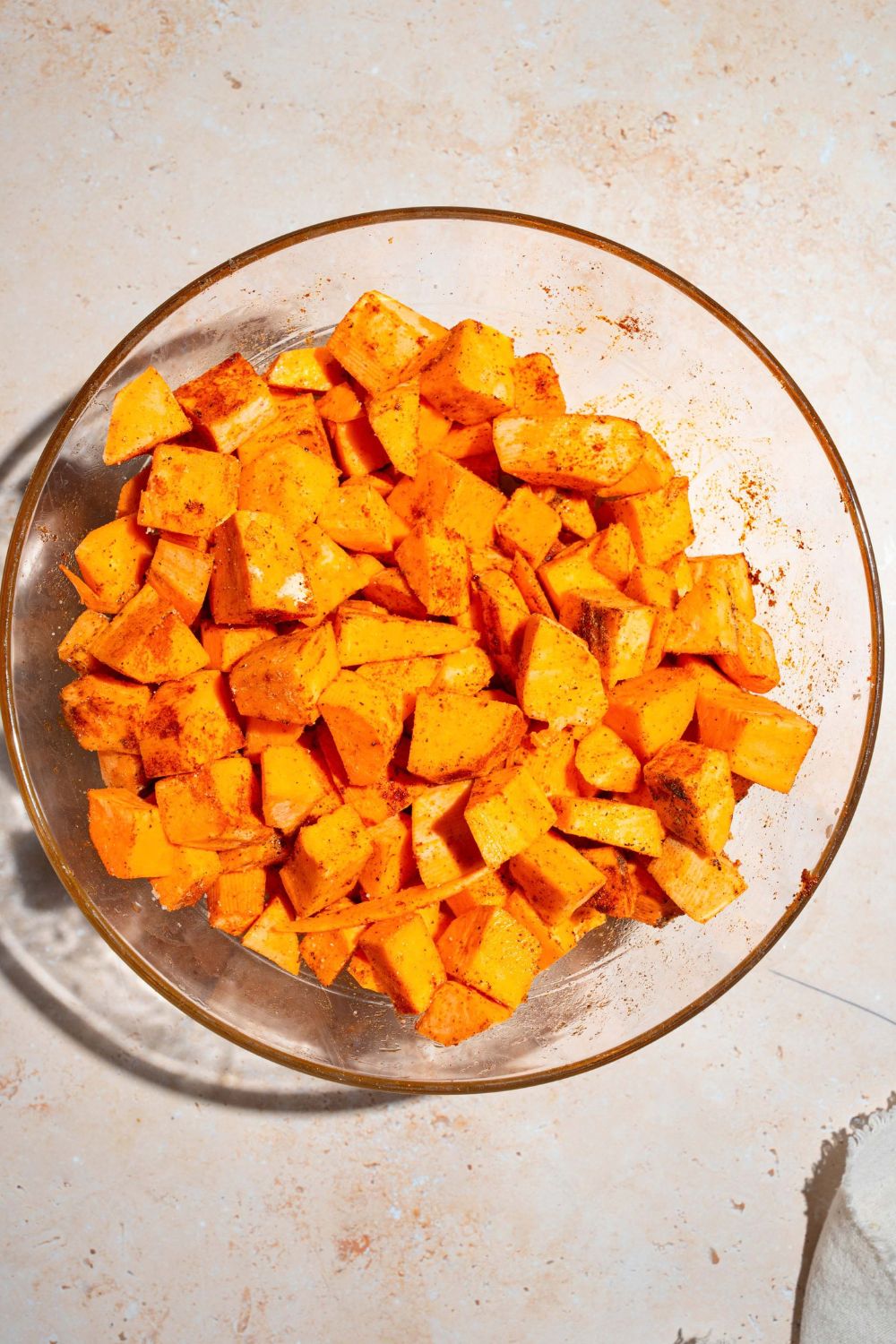 A large bowl with cubed sweet potatoes tossed in seasonings. The bowl is on a tan counter.