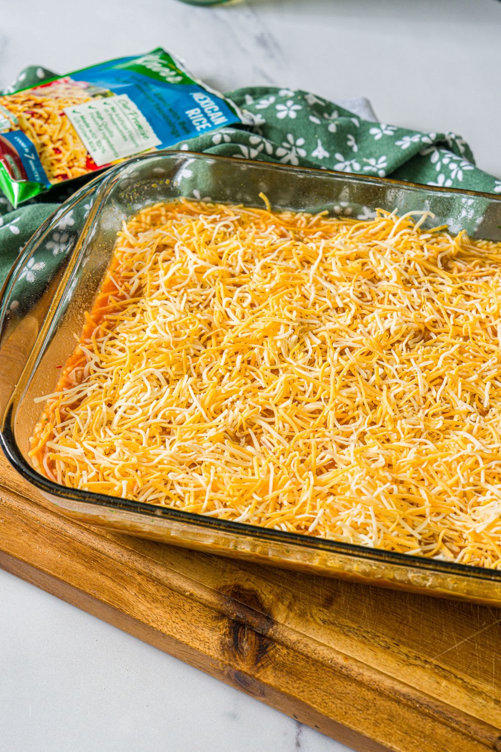 A baking dish with a swim chicken and rice mixture topped with shredded cheese. The dish is on a wooden board on a marble counter.