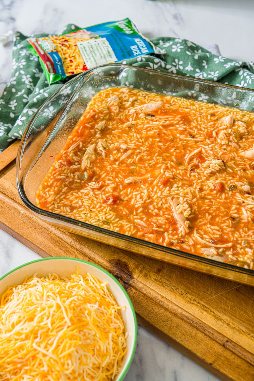 A baking dish with swim chicken and rice mixture. The dish is on a wooden board on a marble counter with a bowl of shredded cheese.