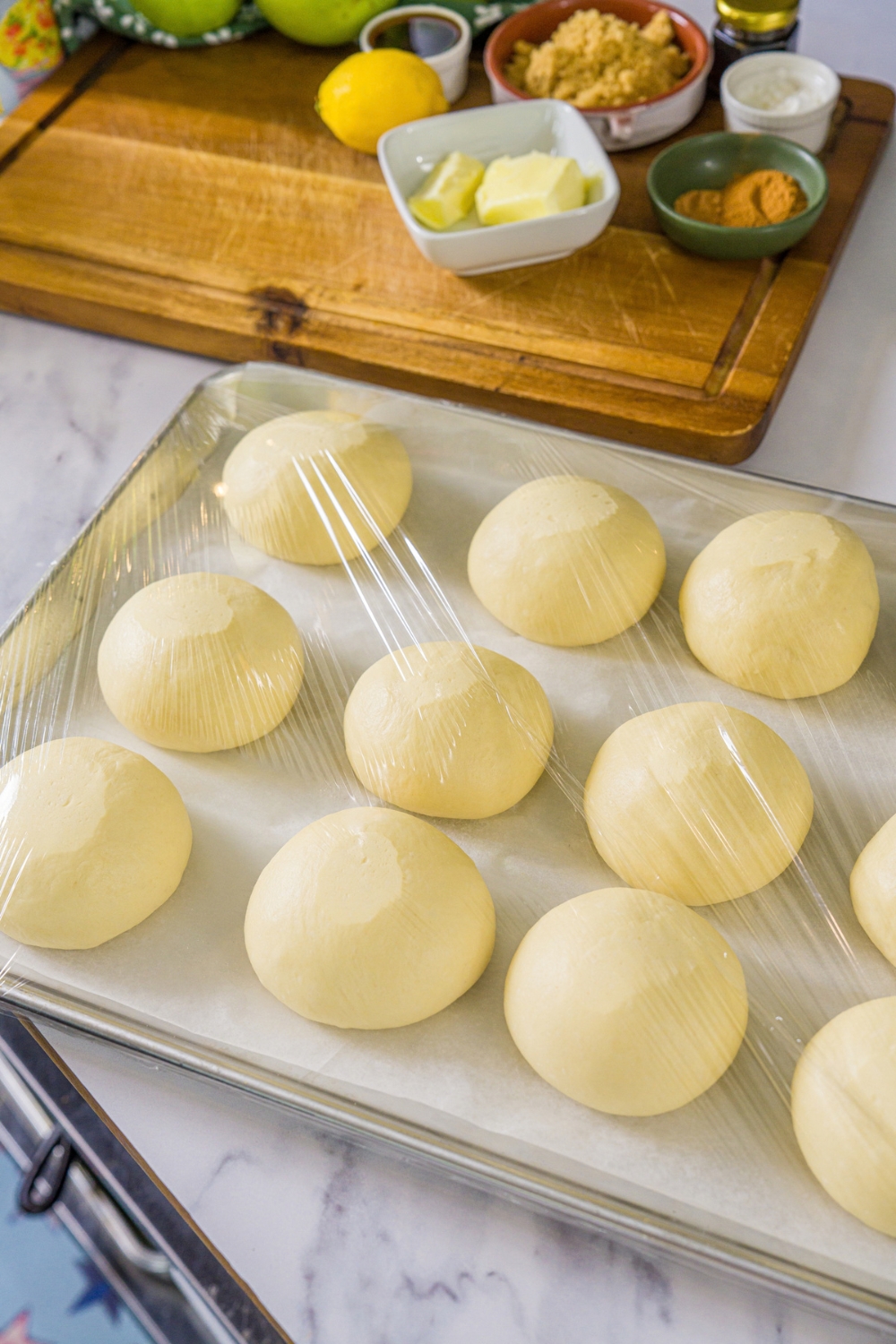 A baking sheet lined with parchment paper with several dough balls. The sheet is covered with plastic wrap.