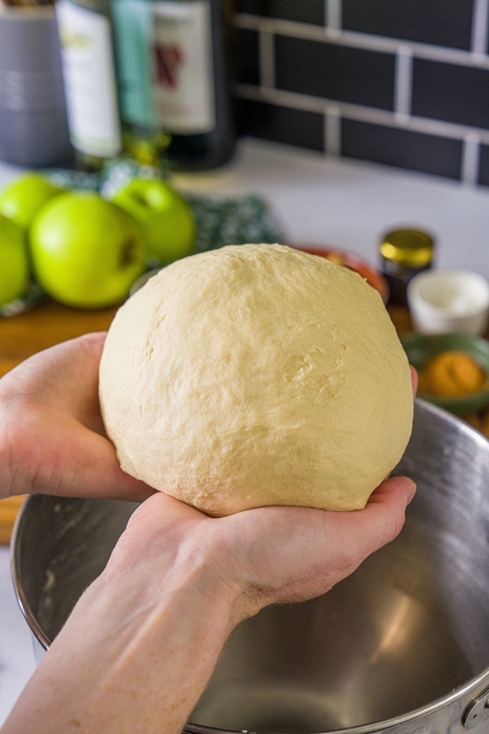 A pair of hands holding a large ball of dough. There is a large mixing bowl on a marble counter blurred in the background.
