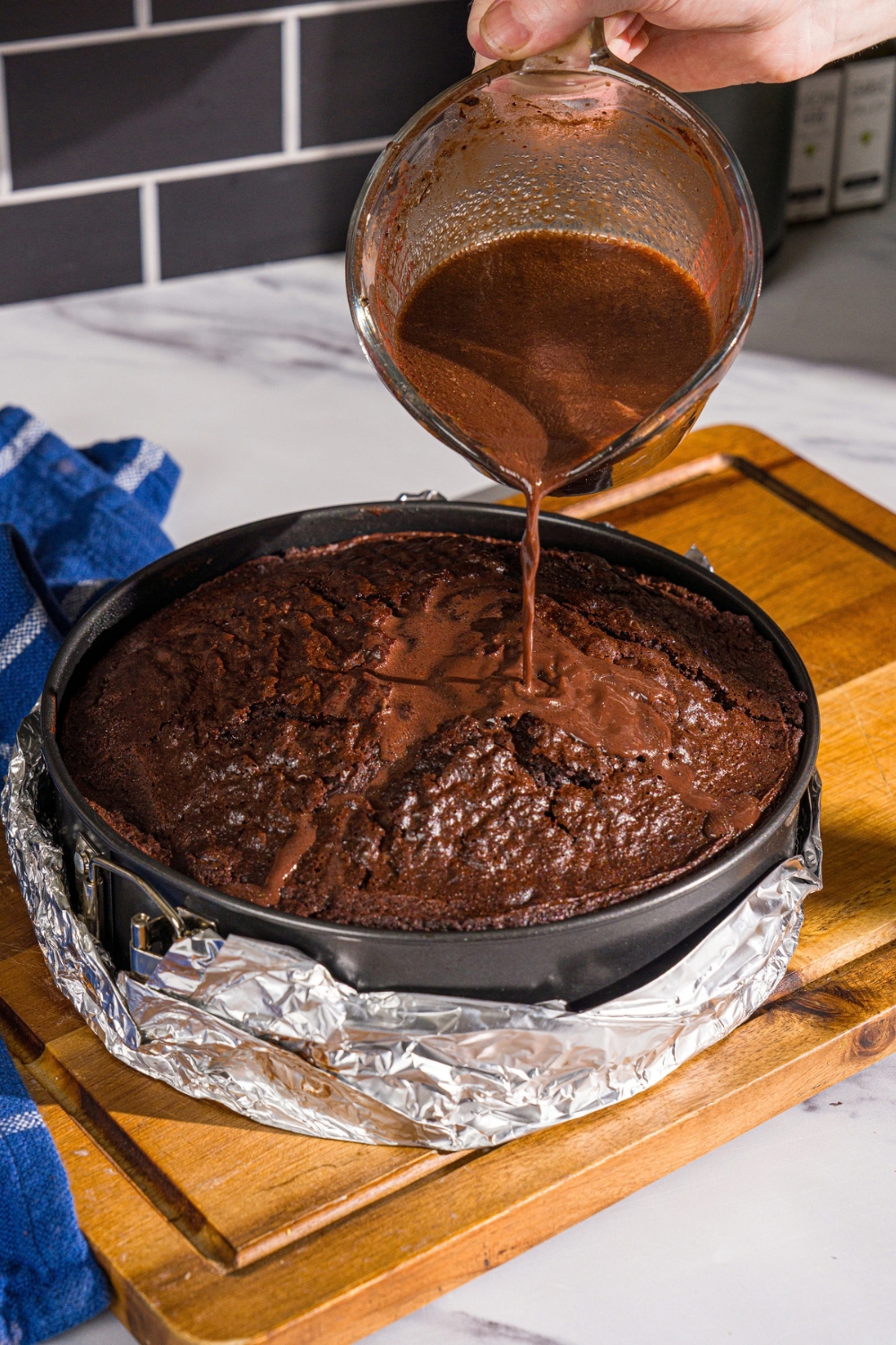 A cake pan wrapped with foil with baked Matilda cake. A measuring cup is pouring a warm soak into the cake. The cake pan is on a wooden board.