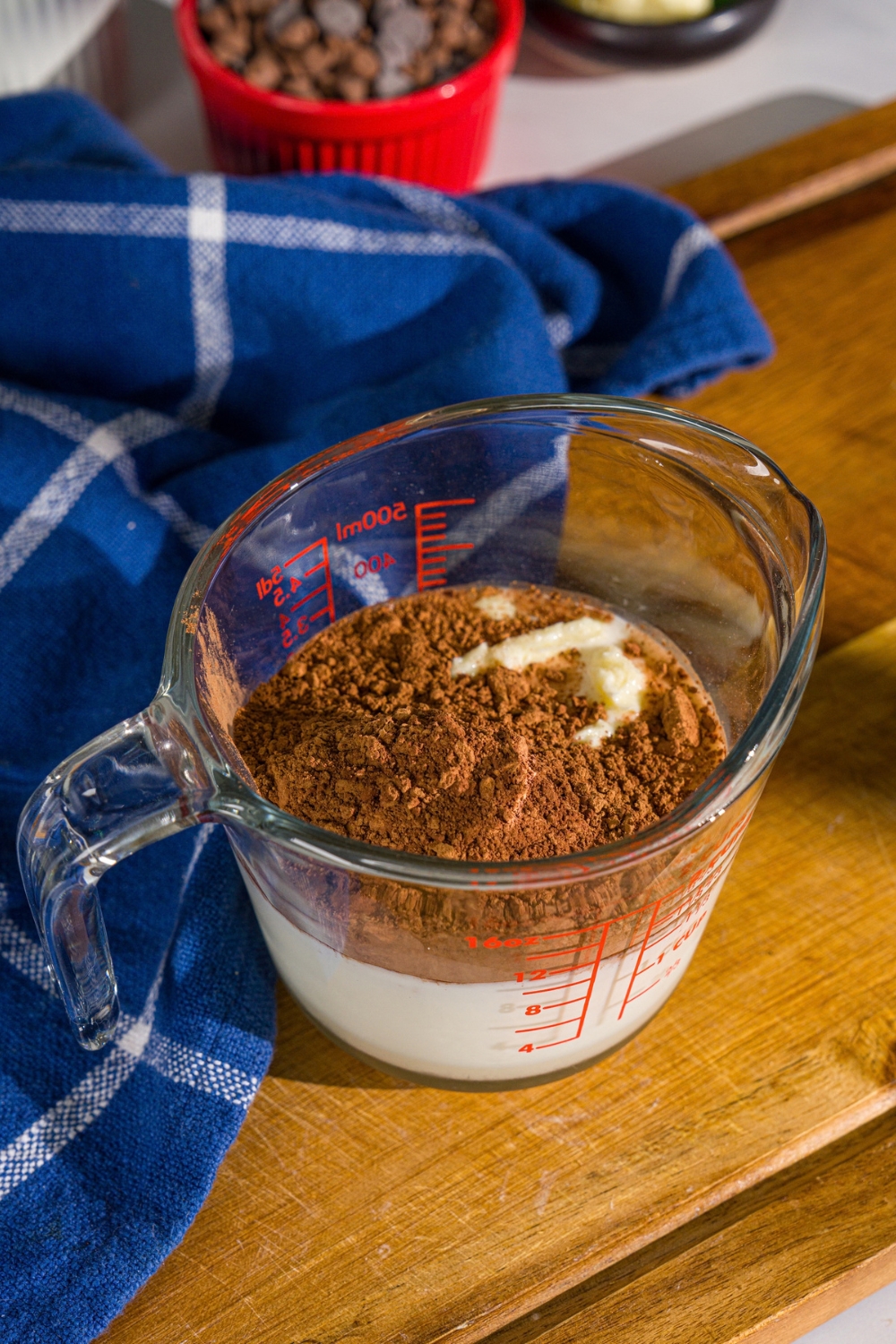 A measuring cup with milk, sugar, cocoa powder, and butter. The cup is on a wooden board with a blue cloth napkin.