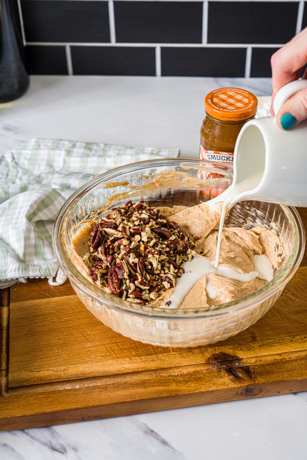 A hand pouring cream into a bowl of cinnamon roll dip that is filled with pecans.