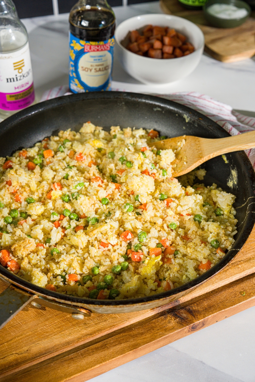 A skillet with a fried rice mixture including egg, peas, and carrots. A wooden spoon is stirring the rice. The skillet is on a wooden board.