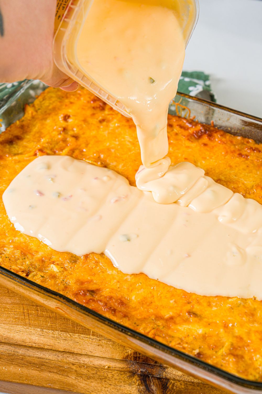 A baking dish with baked swim chicken and rice with a bowl of queso being poured on top. The dish is on a wooden board,