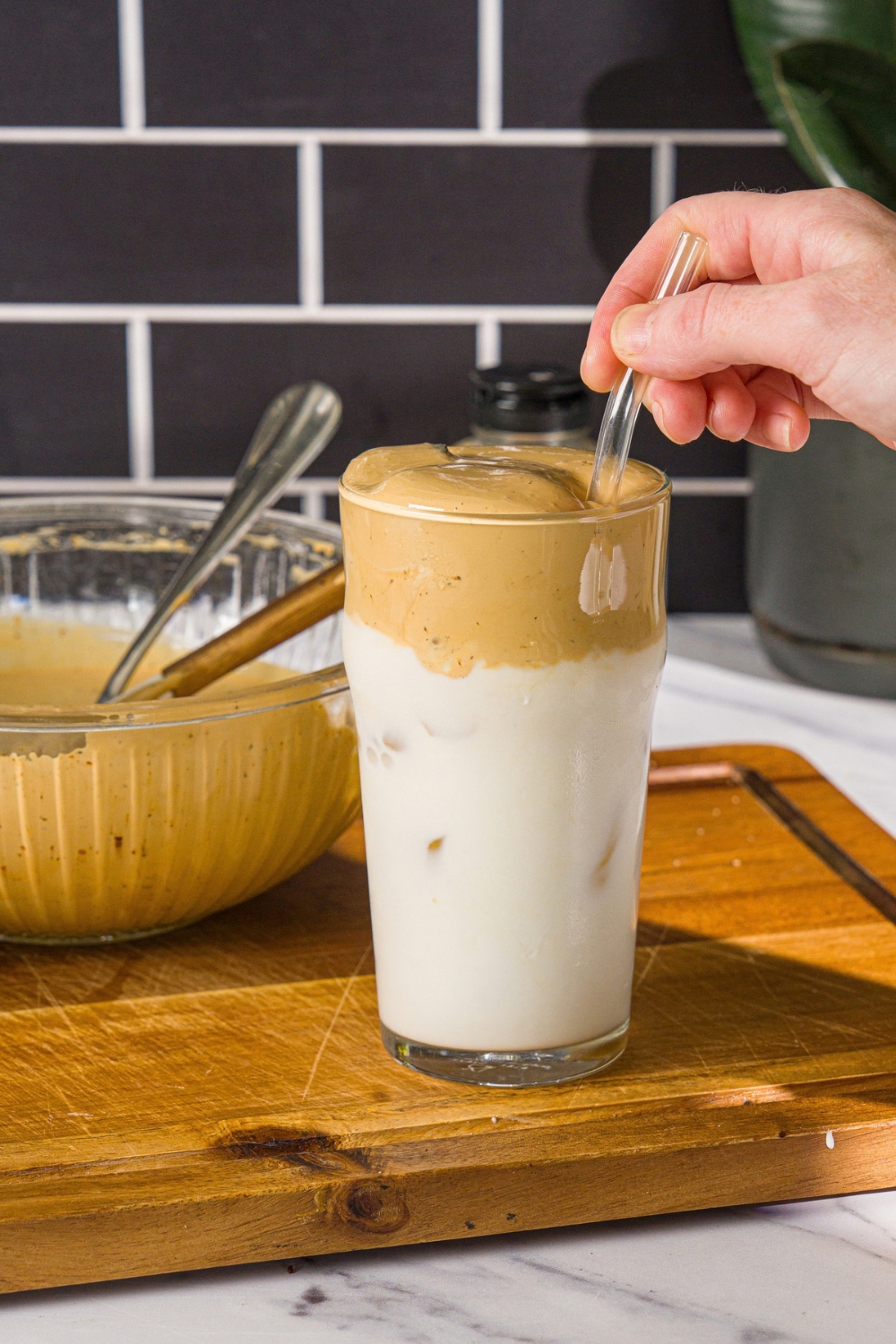A glass with iced milk topped with whipped honey coffee. A straw is stirring the coffee. The glass is on a wooden board with a bowl of honey whipped coffee.
