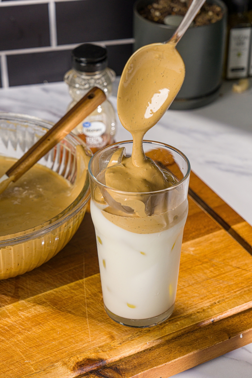 A glass of iced milk with whipped honey coffee being spooned into the glass. The glass is on a wooden board with a bowl of whipped honey coffee.