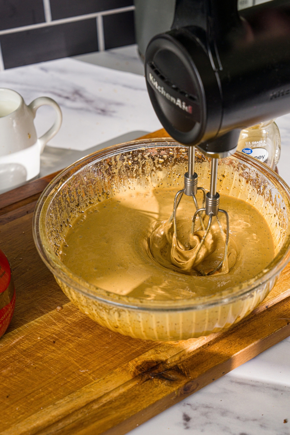 A glass bowl with a hand mixer mixing whipped honey coffee mixture. The bowl is on a wooden board.