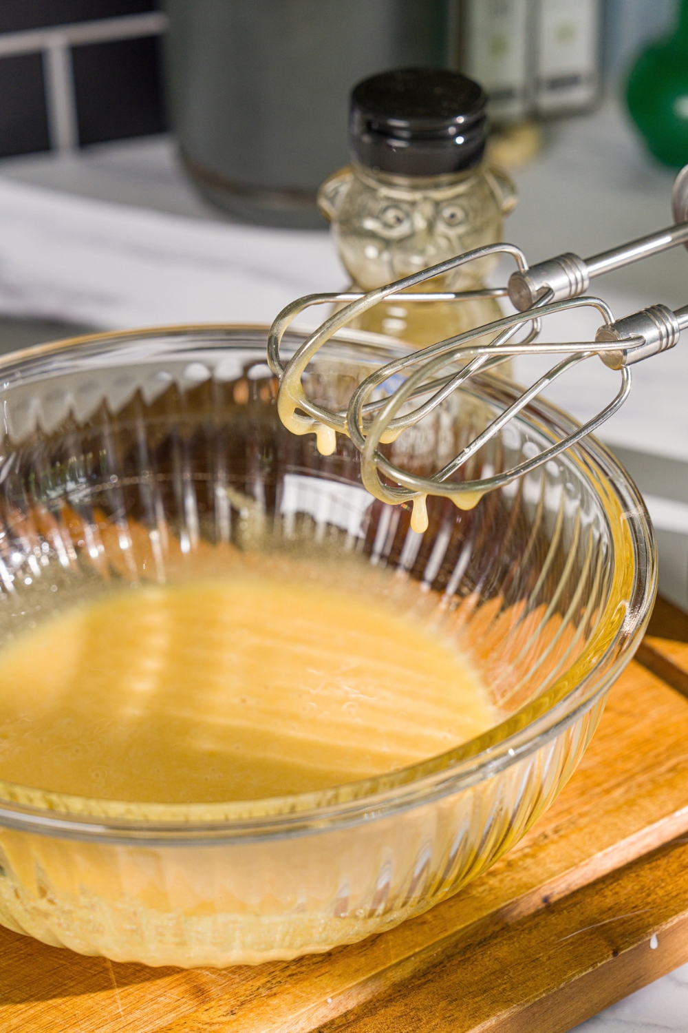 A glass bowl with a mixer dipped in a whipped honey. The bowl is on a wooden board.