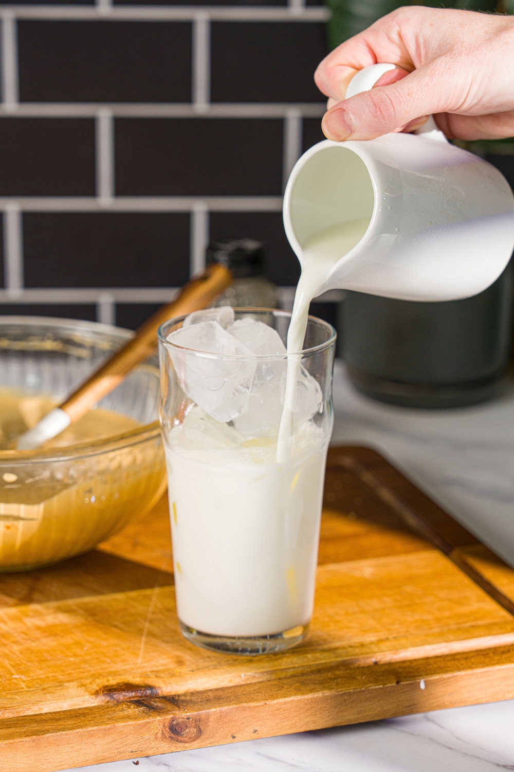 A glass of ice with milk being poured into the glass. The glass is on a wooden board with a bowl of whipped honey coffee.