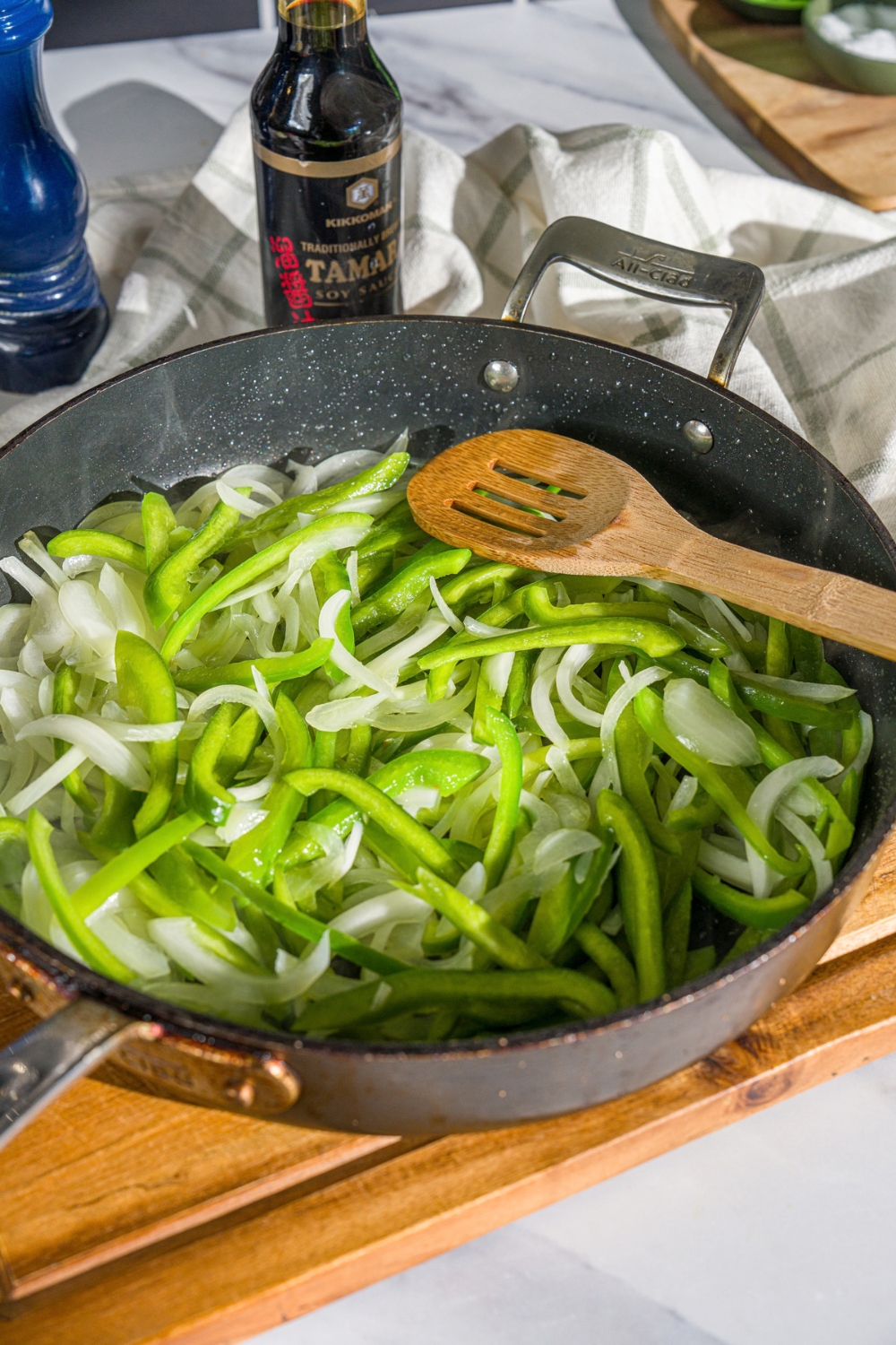A skillet with sliced green peppers and onions. There is a wooden spoon in the skillet. The skillet is on a wooden board.