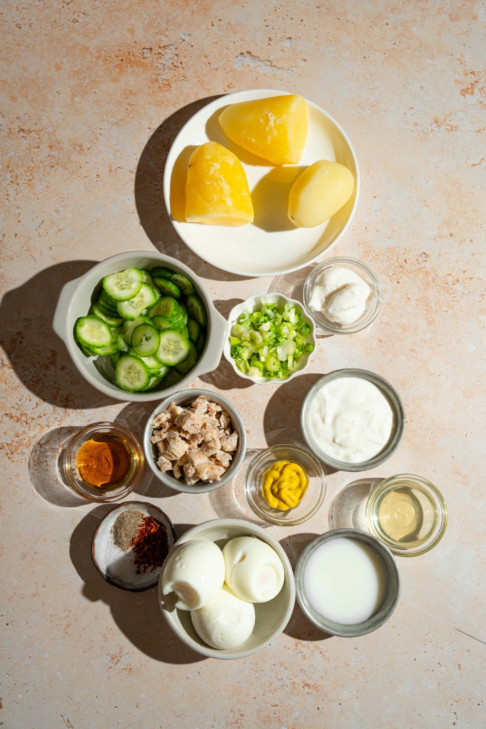 A tan counter with several bowls containing ingredients to make Japanese potato salad including potatoes, hard boiled eggs, cucumbers, chicken, green onion, yogurt, mayo, mustard, honey, and more.