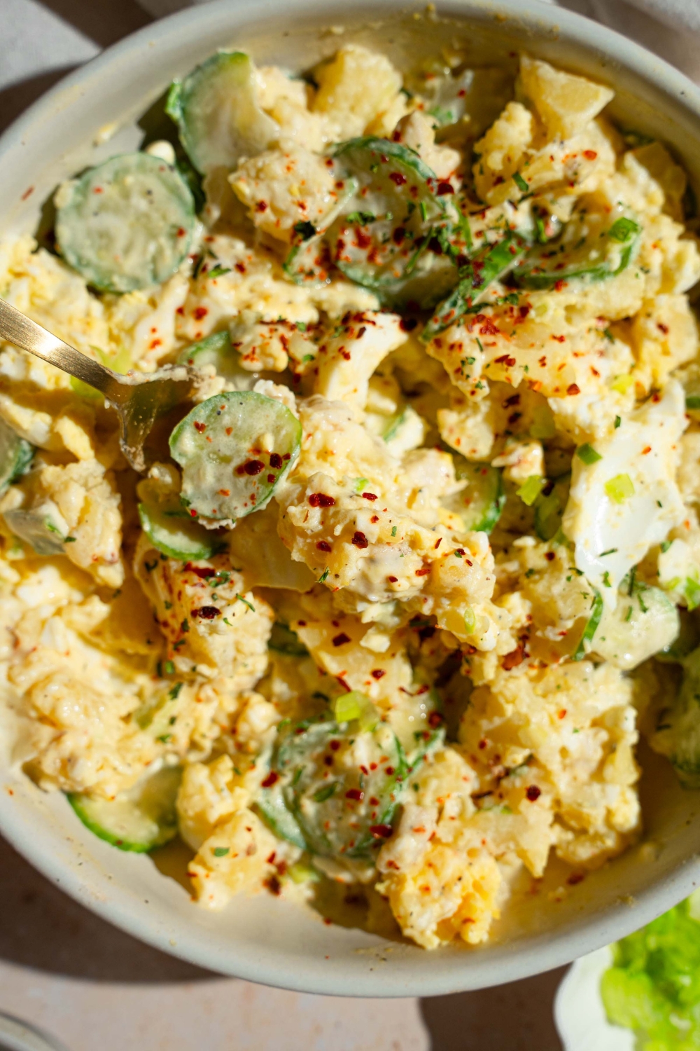 A white bowl with Japanese potato salad with potatoes, eggs, chicken, cucumbers, and green onion tossed in a creamy dressing and topped with seasonings. There is a fork in the bowl.