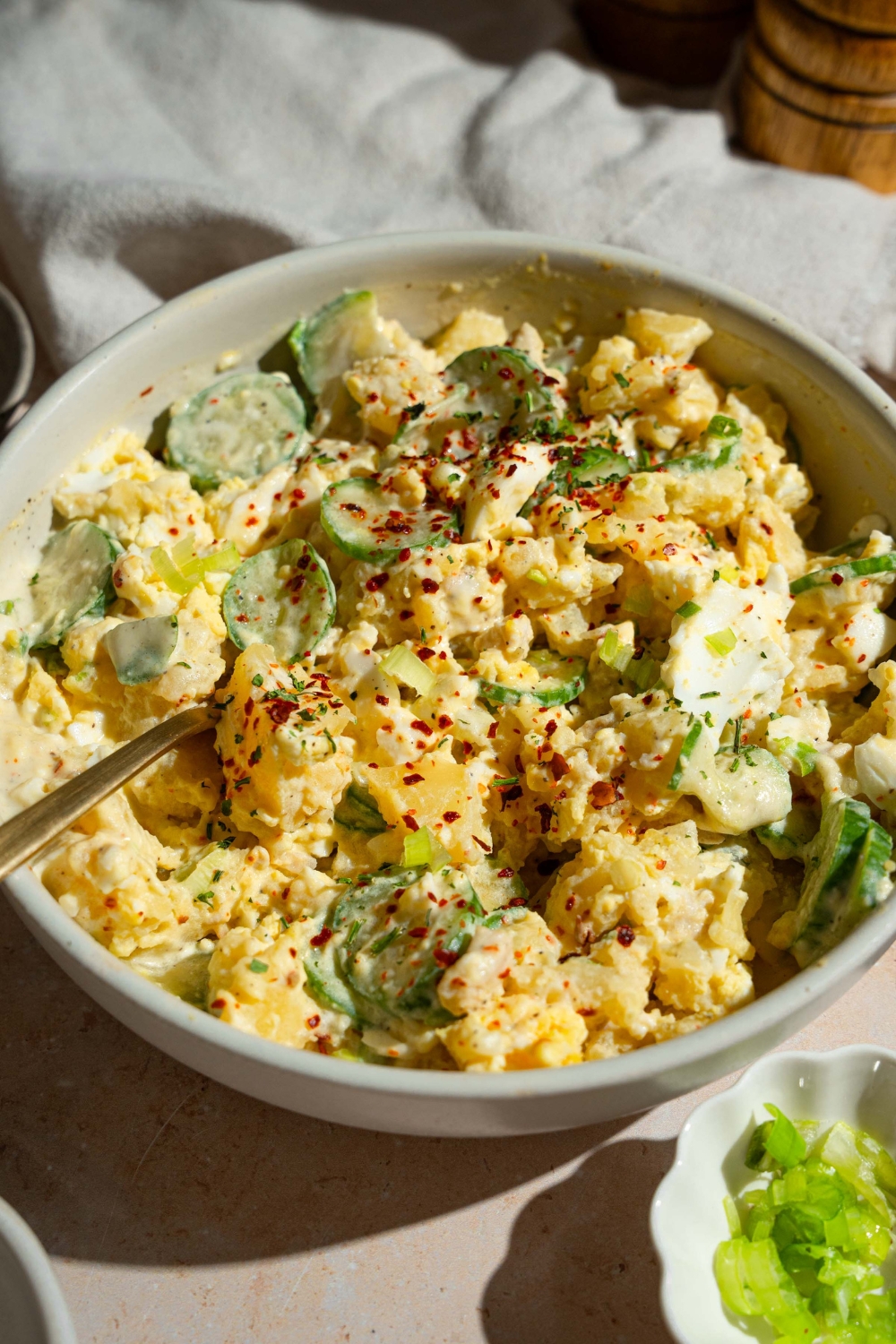 A white bowl with Japanese potato salad with potatoes, eggs, chicken, cucumbers, and green onion tossed in a creamy dressing and topped with seasonings. There is a fork in the bowl. The bowl is on a tan counter with a small bowl of garnishes.