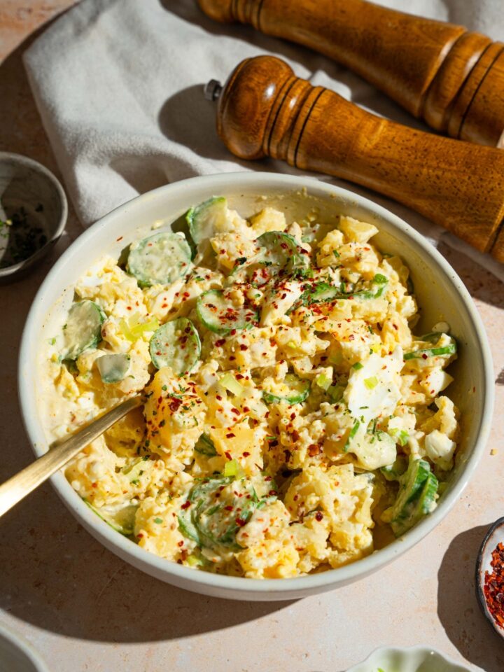 A white bowl with Japanese potato salad with potatoes, eggs, chicken, cucumbers, and green onion tossed in a creamy dressing and topped with seasonings. There is a fork in the bowl. The bowl is on a tan counter with a salt and pepper mill and a white cloth napkin.