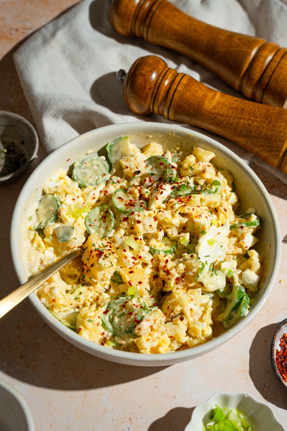 A white bowl with Japanese potato salad with potatoes, eggs, chicken, cucumbers, and green onion tossed in a creamy dressing and topped with seasonings. There is a fork in the bowl. The bowl is on a tan counter with a salt and pepper mill and a white cloth napkin.