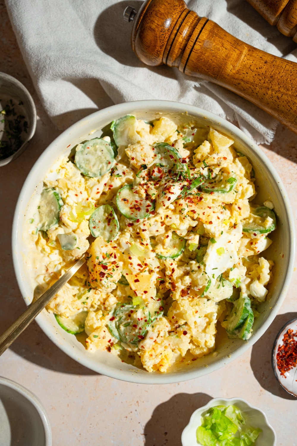 A white bowl with Japanese potato salad with potatoes, eggs, chicken, cucumbers, and green onion tossed in a creamy dressing and topped with seasonings. There is a fork in the bowl. The bowl is on a tan counter with small bowls of garnishes and a salt and pepper mill.