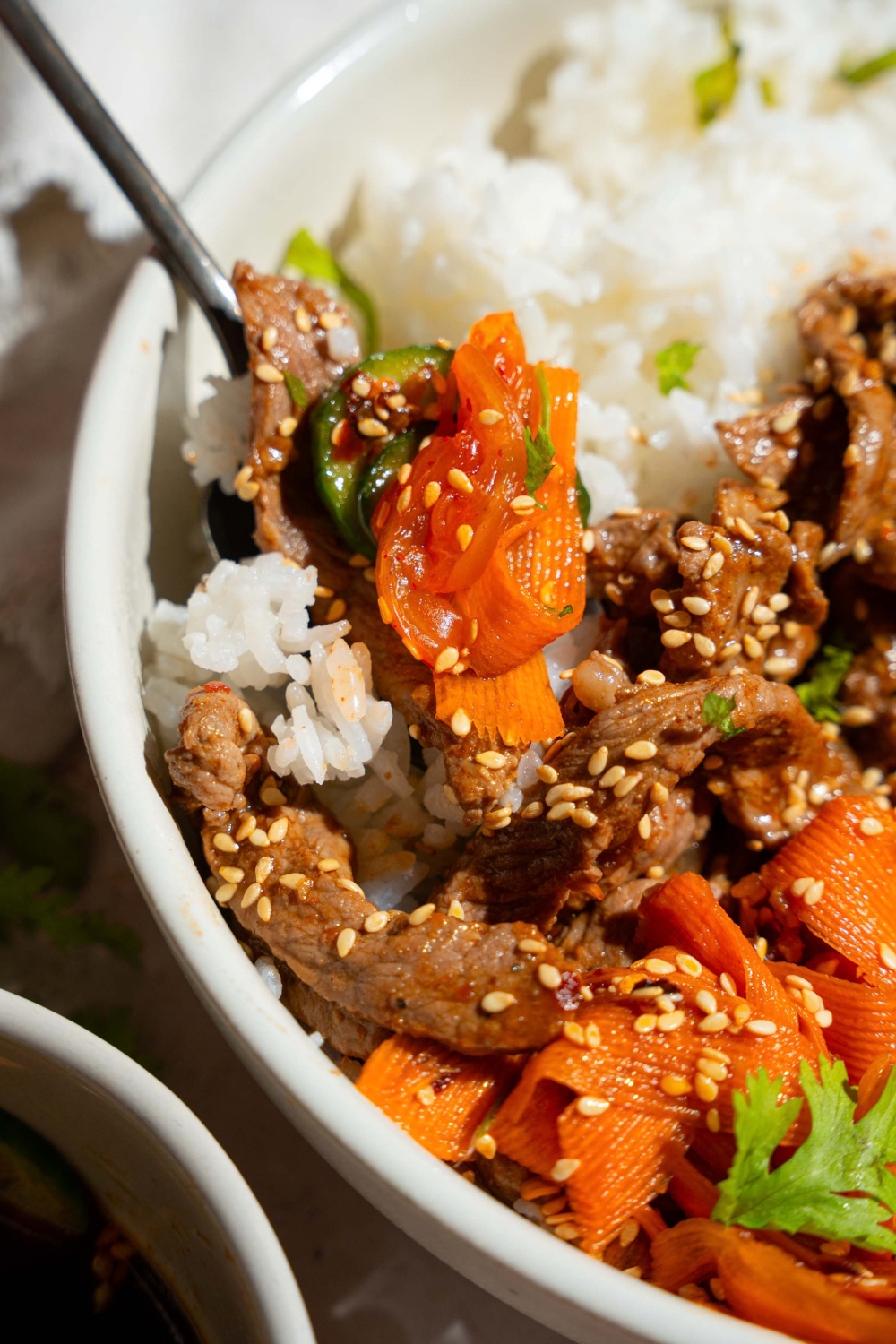 A white bowl with a beef bulgogi bowl with rice, beef bulgogi, and a cucumber carrot salad, all garnished with sesame seeds. A fork is taking a bite from the bowl.