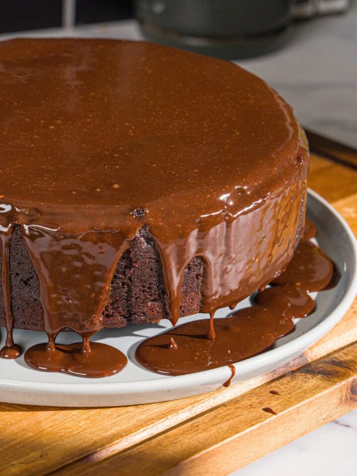 A white plate with a whole Matilda cake covered with chocolate ganache. The plate is on a wooden board.