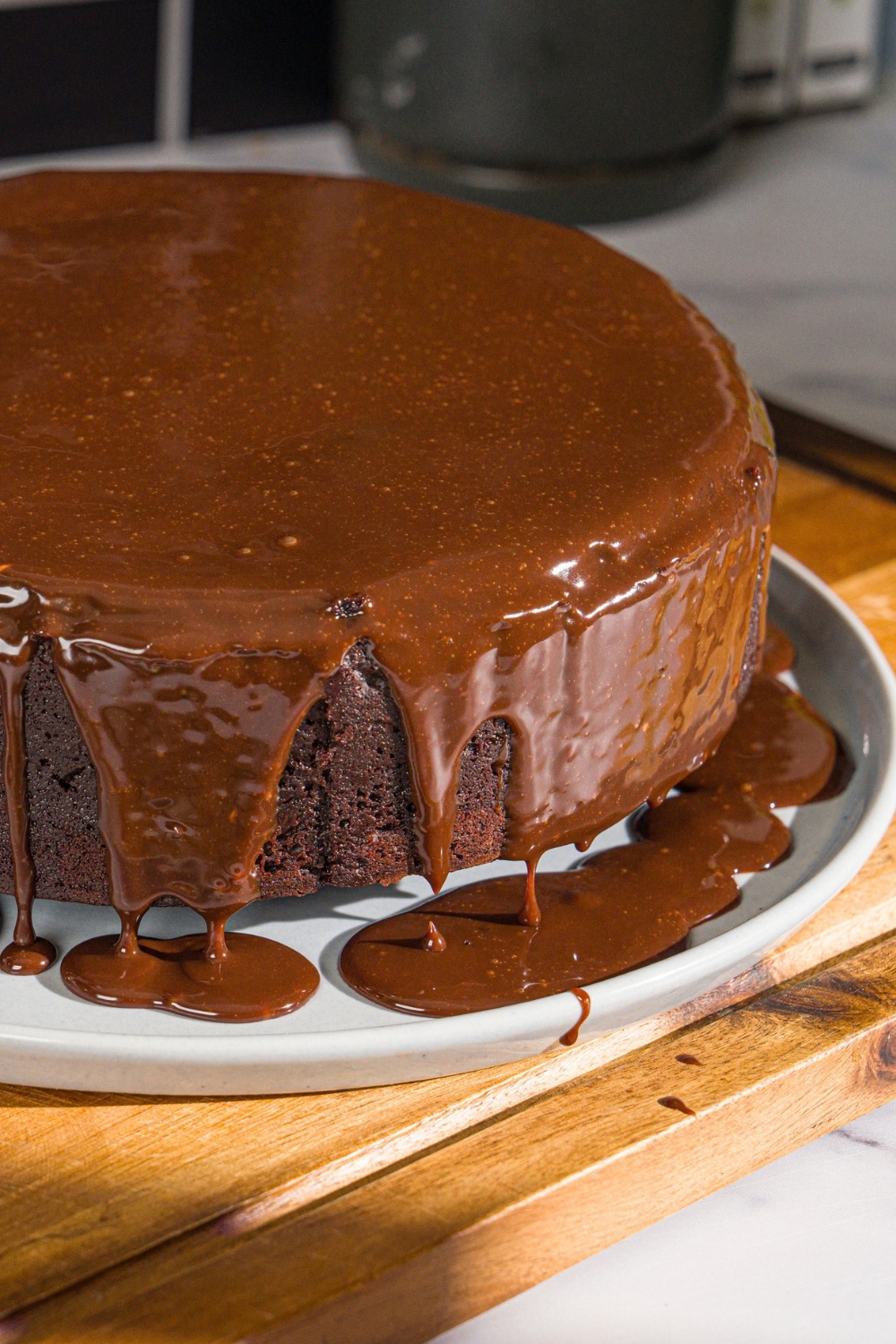 A white plate with a whole Matilda cake covered with chocolate ganache. The plate is on a wooden board.