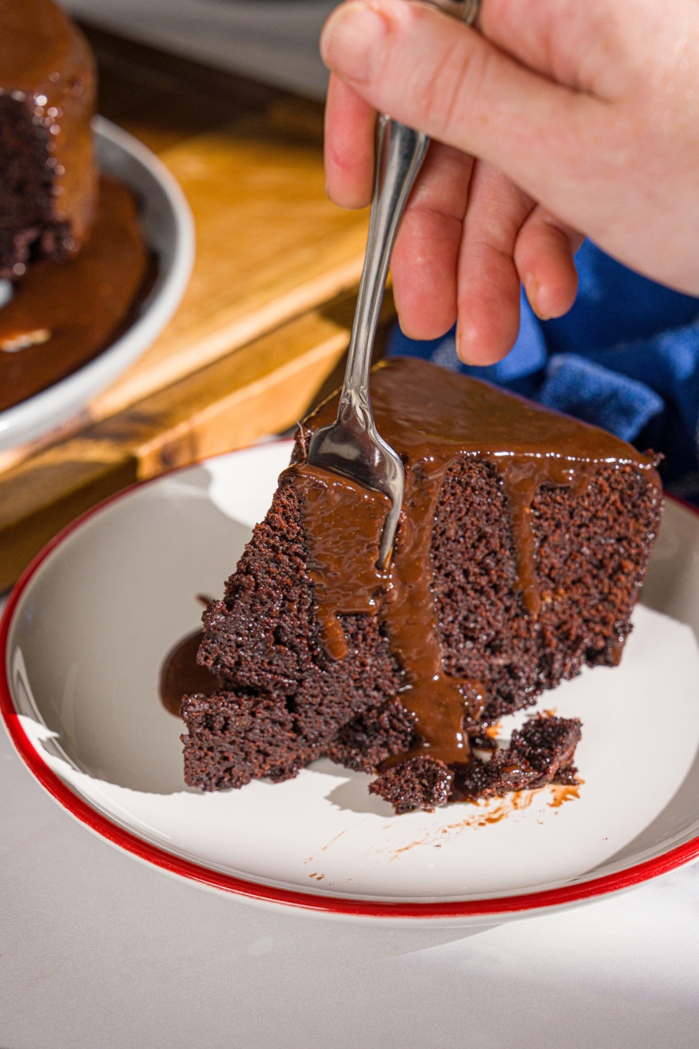 A white plate with a slice of Matilda cake topped with chocolate ganache. A fork is taking a bite from the cake.