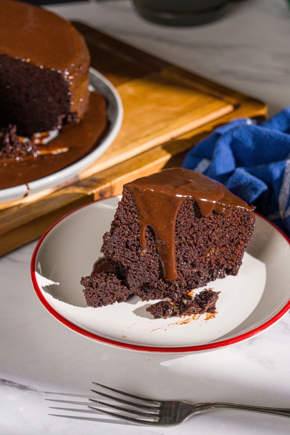 A white plate with a slice of Matilda cake topped with chocolate ganache. The plate is on a marble counter with a wooden board with a plate of cake.