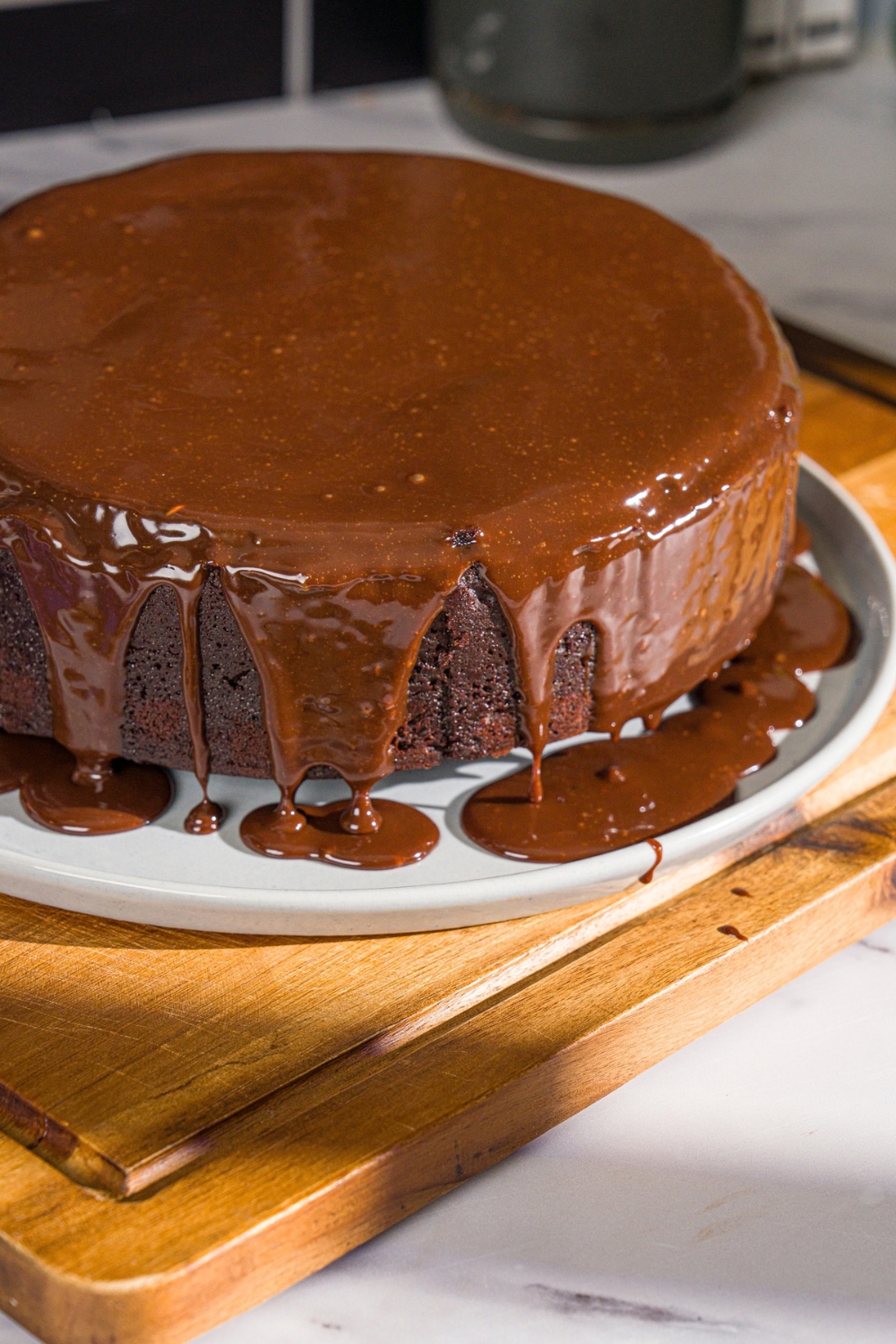 A white plate with a whole Matilda cake covered with chocolate ganache. The plate is on a wooden board.